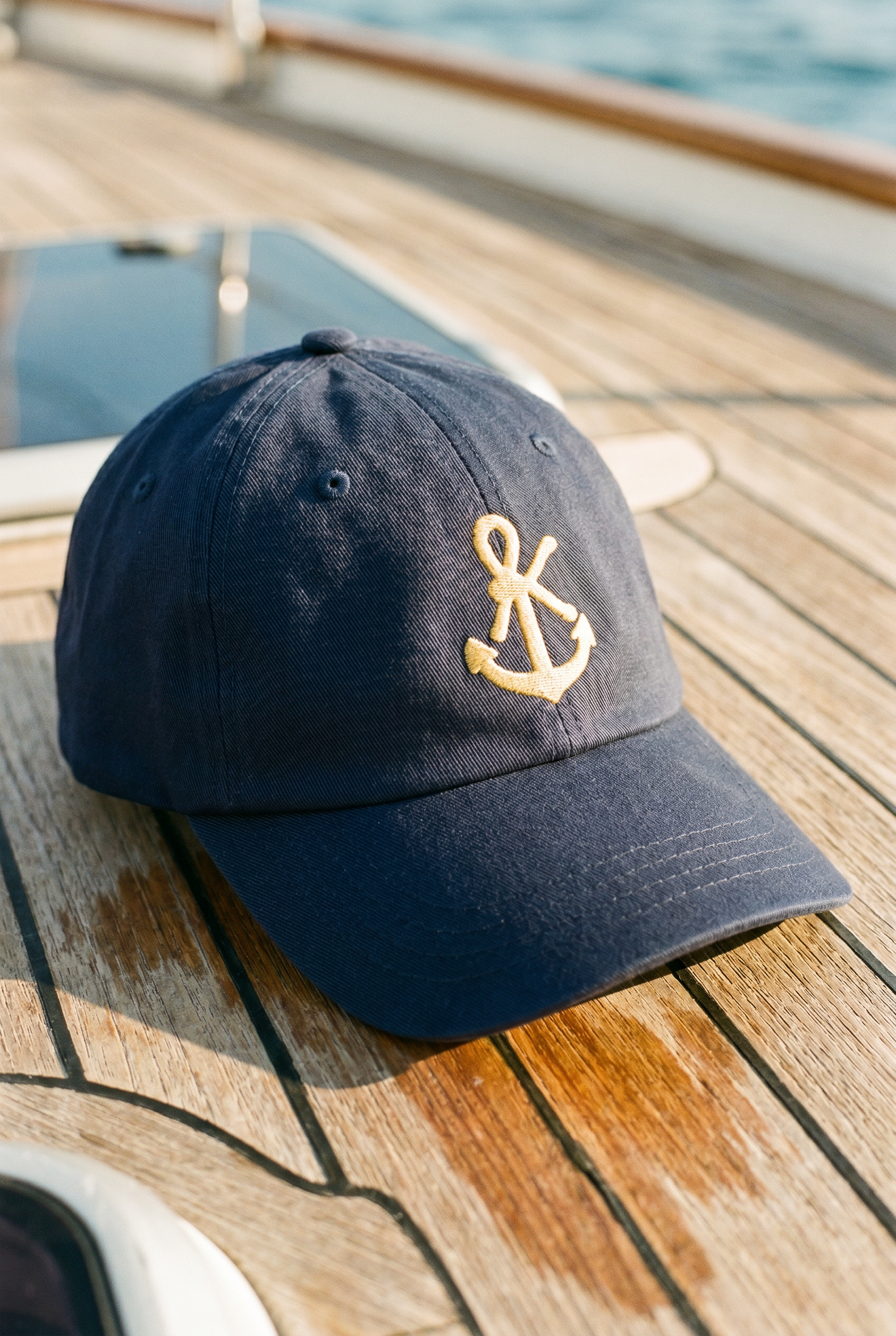 SŁOŃCE Signature Baseball Cap Deep Baltic mit goldener Stickerei auf einem Holzdeck eines Segelbootes.